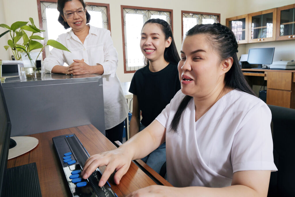 A blind Asian woman works at a computer, with her two female Asian colleagues smiling behind her