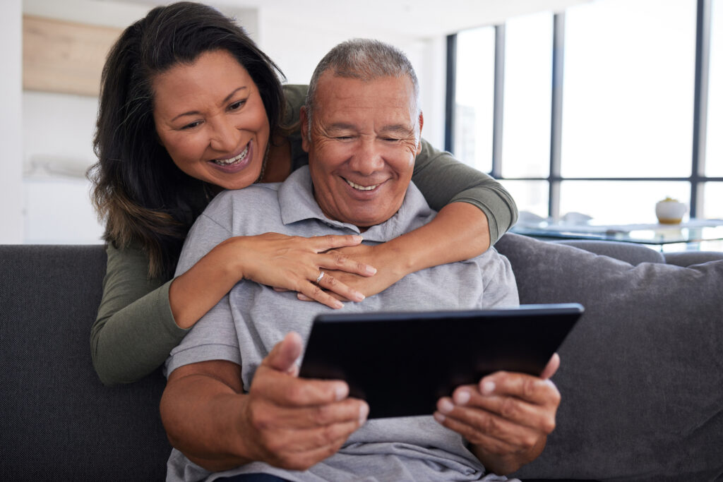Latino senior married couple sit on couch and smile at tablet device