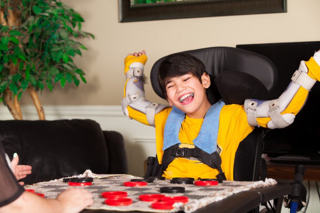 Young mixed-race boy with cerebral palsy sits in his wheelchair with arms raised and a smile on his face.
