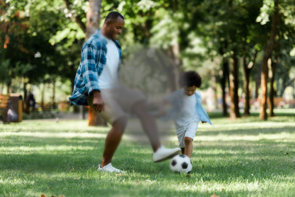 A young Black father and son play with a football outside in a sunny park. There is a dark blurred spot in the middle of the image, to represent the effect of macular degeneration edited