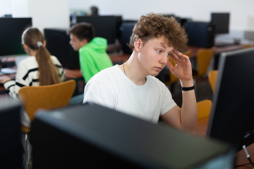 In a college computer lab, a frustrated student in a white tee shirt strains to drown out other people's conversation and focus on his assignment
