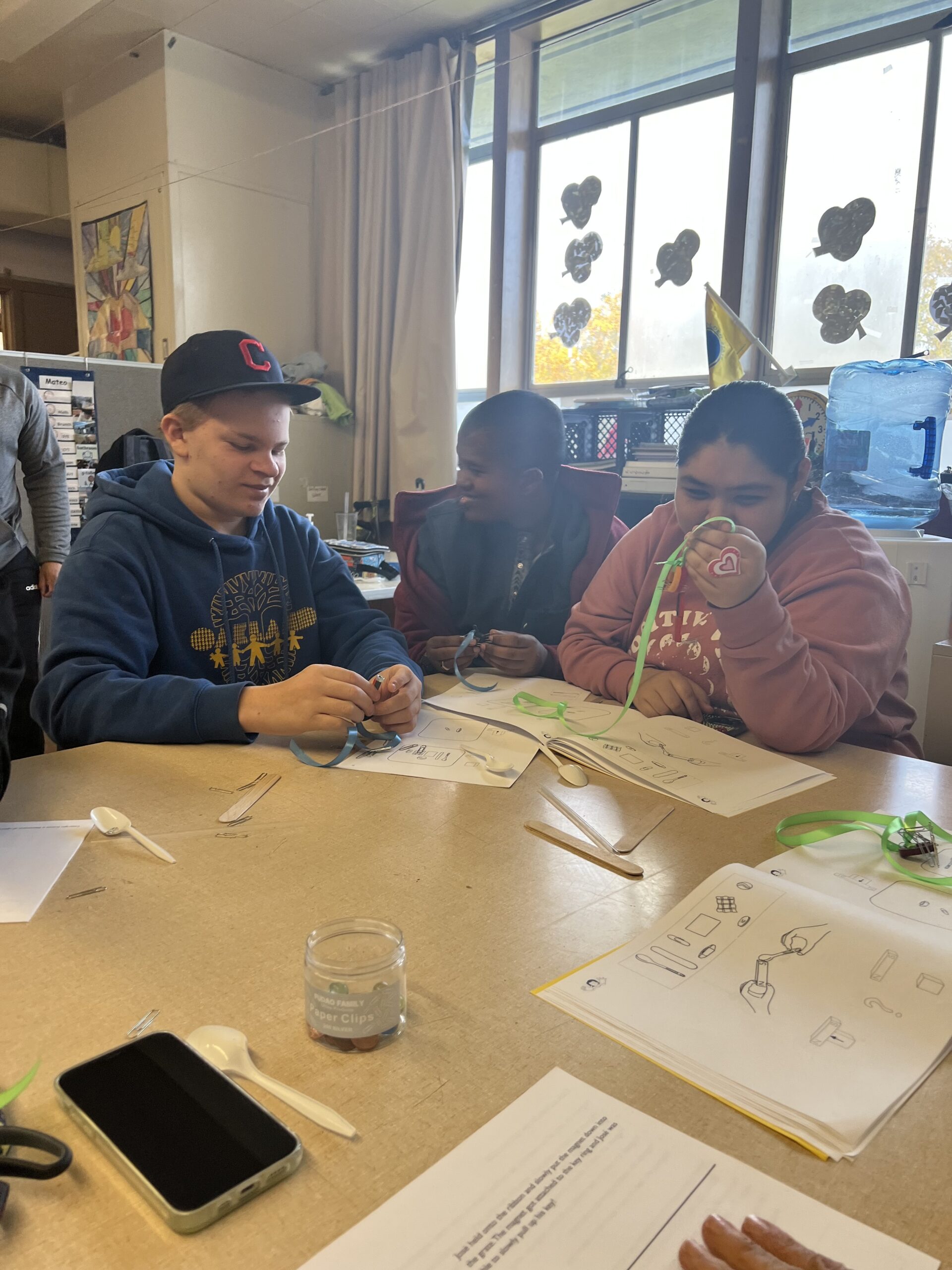 Three high school students sit at a wide tan table in a brightly-lit classroom, smiling while they work together on a hands-on science experiment involving magnets, paper clips, and brightly colored ribbons.