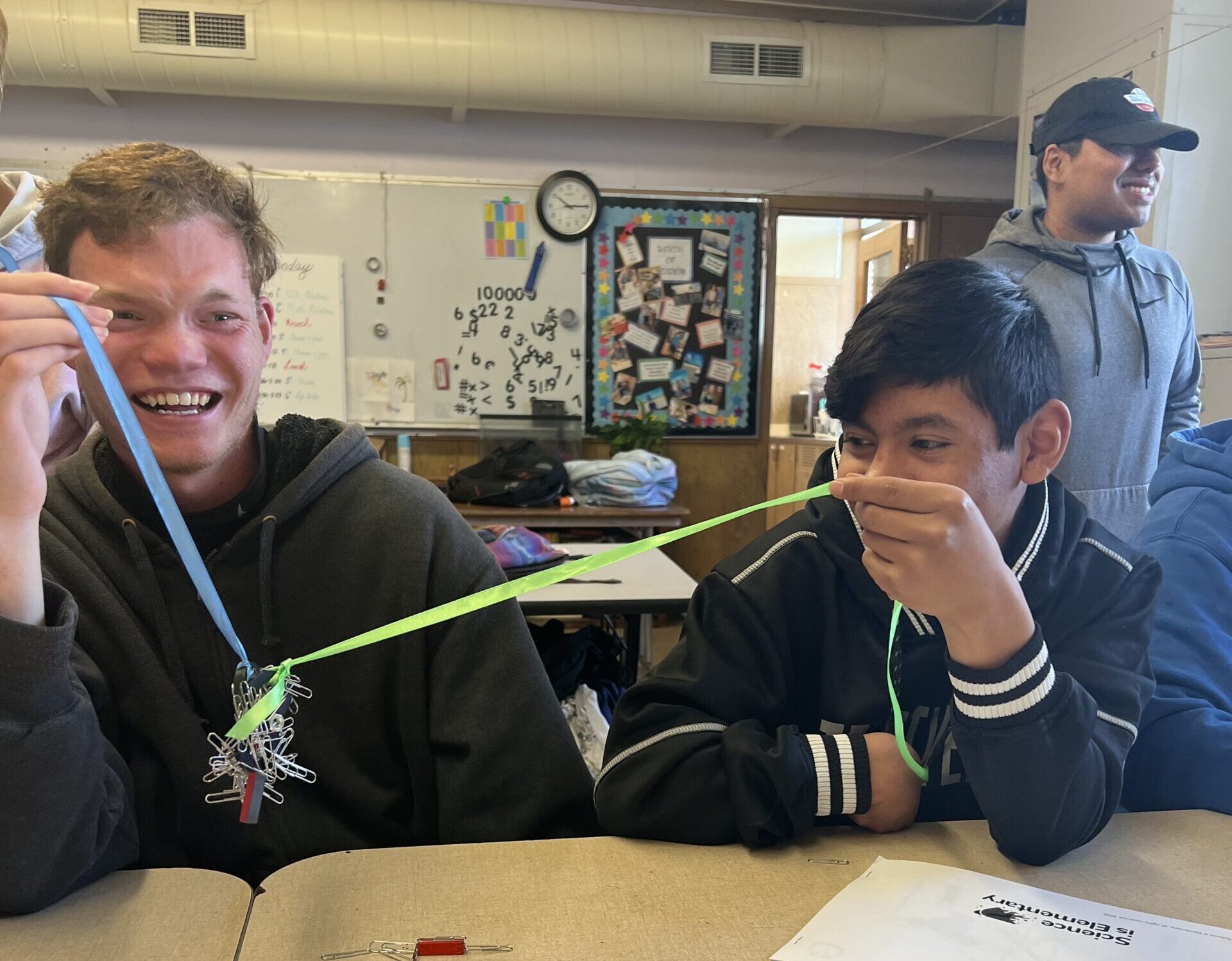 Two high school boys sit in a crowded classroom, laughing as they work on an experiment using magnets and ribbons to hold a pile of paperclips in the air between them.