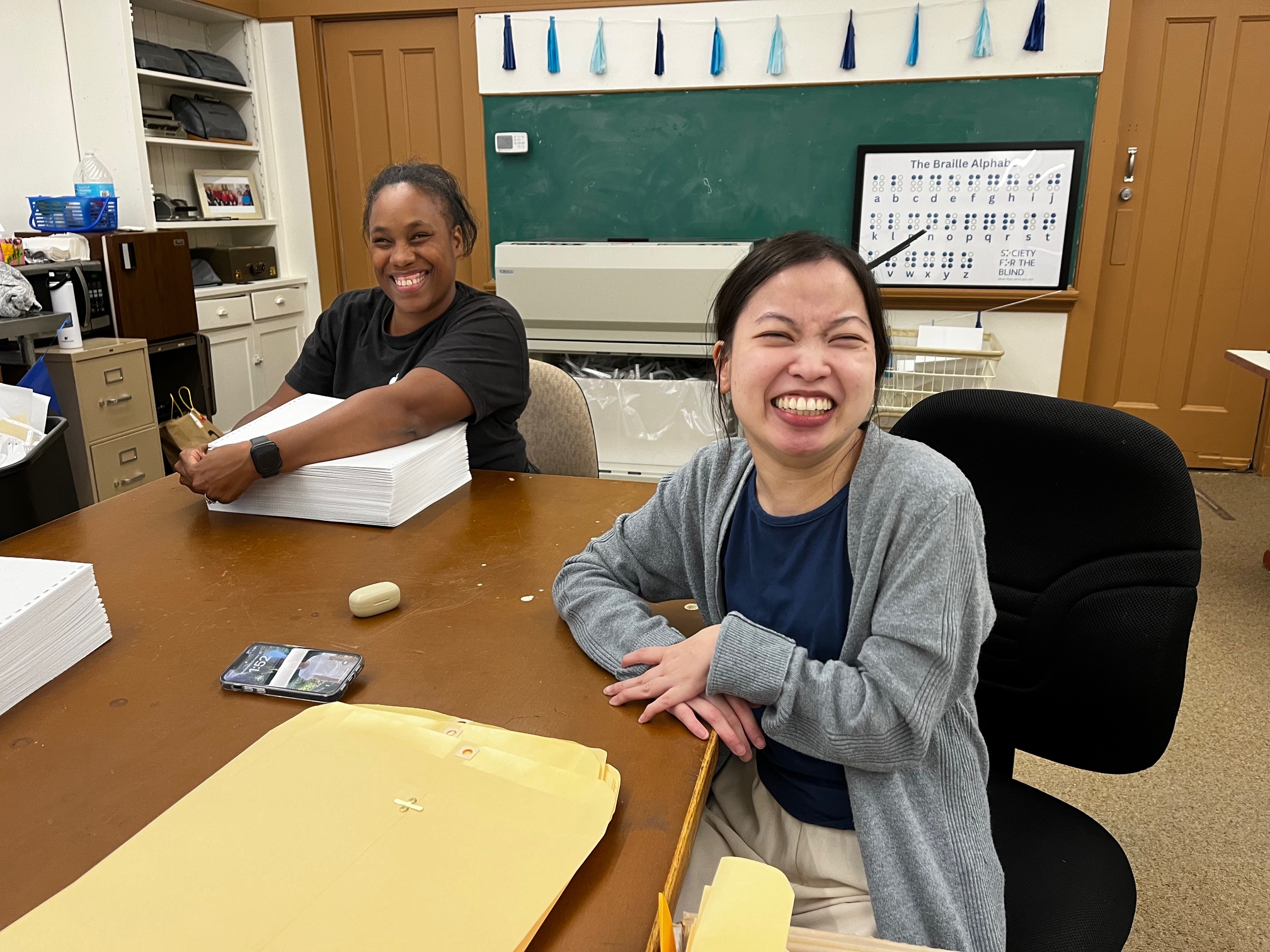 A Black woman wearing a black tee shirt and smart watch and an Asian woman in a grey cardigan take a break from creating Braille materials at a long brown table to smile for the camera.