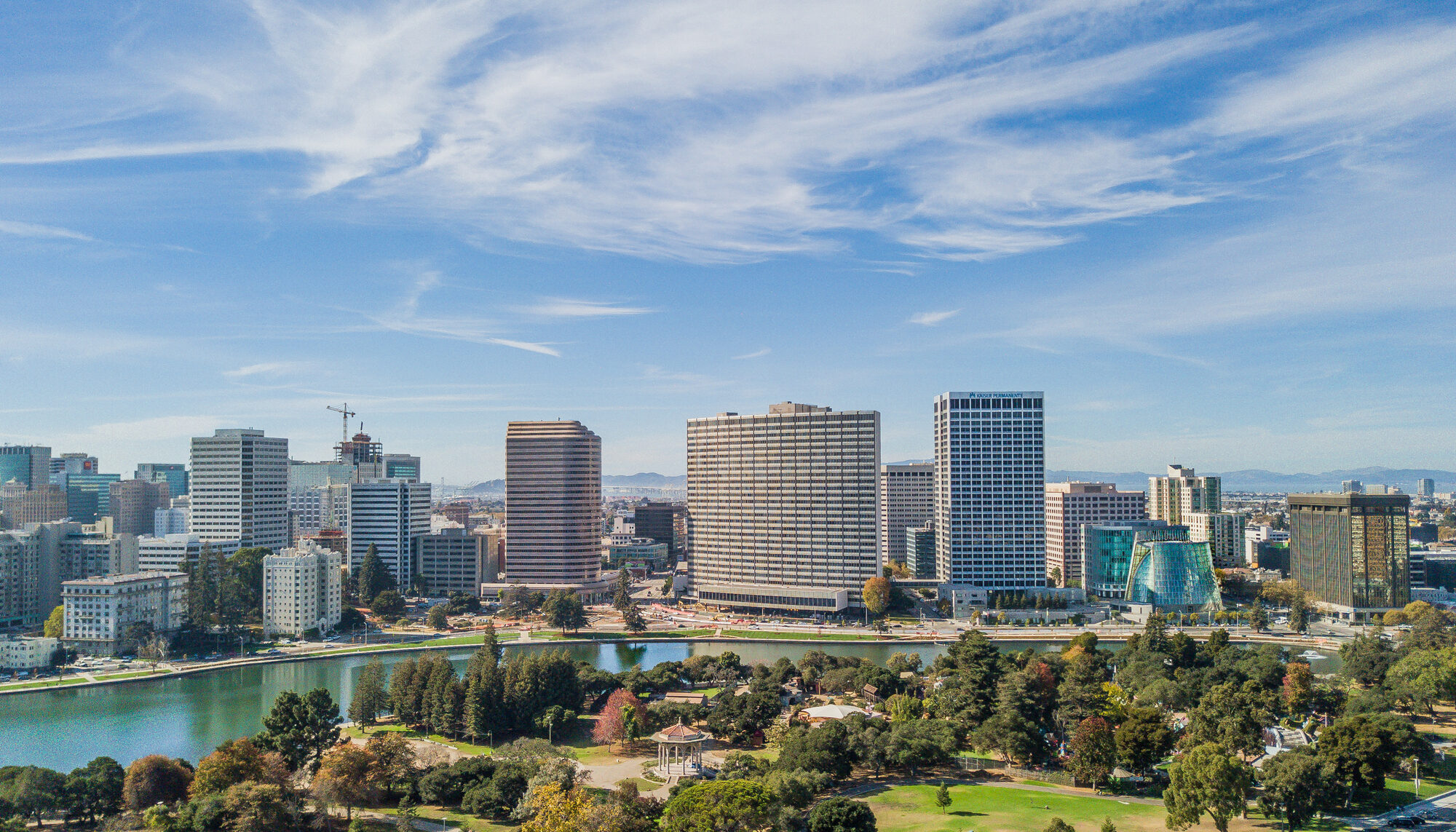 Aerial view of Oakland, California viewed from a tall vantage point above a city park with a pagoda in the middle. A green-blue lake sits in the middle of the photo, with thin clouds in a bright blue sky.