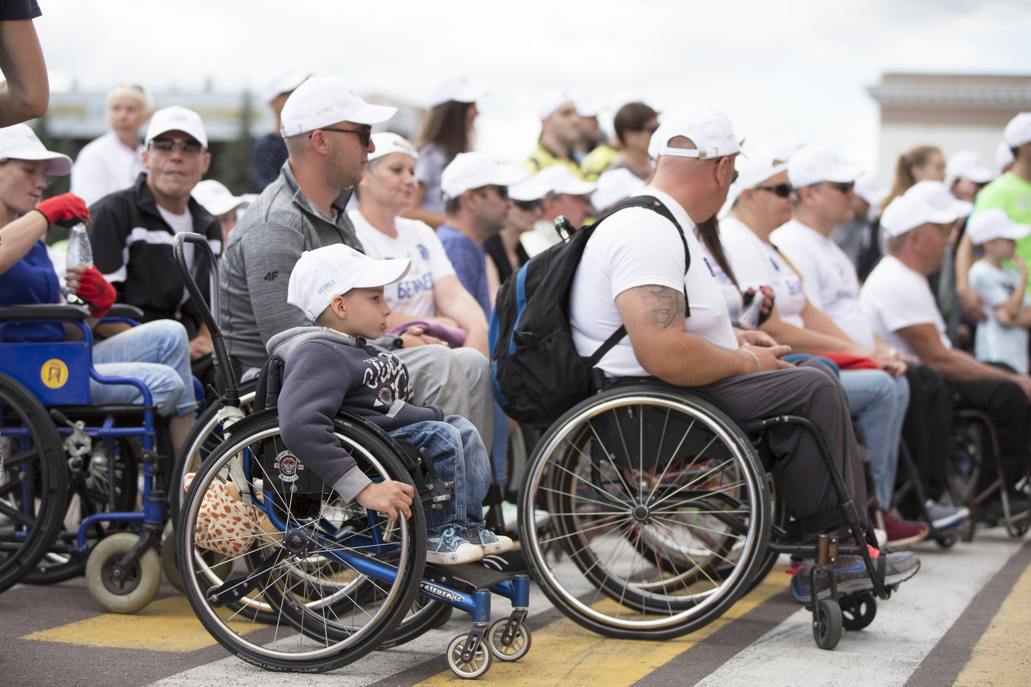 A diverse group of wheelchair users wearing white baseball caps gather for an outdoor charity event. Most are older adults, and there is a young boy in the foreground seated in his wheelchair with a determined expression. 