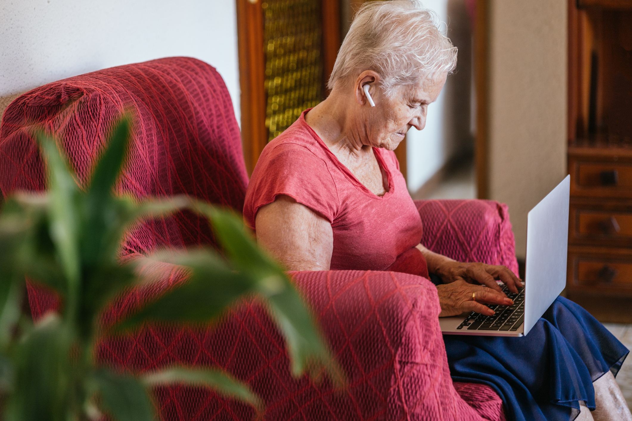 An elderly white woman with short, thin white hair and a red tee shirt sits in a red patterned armchair at home, reading articles on her laptop with white earbuds in.