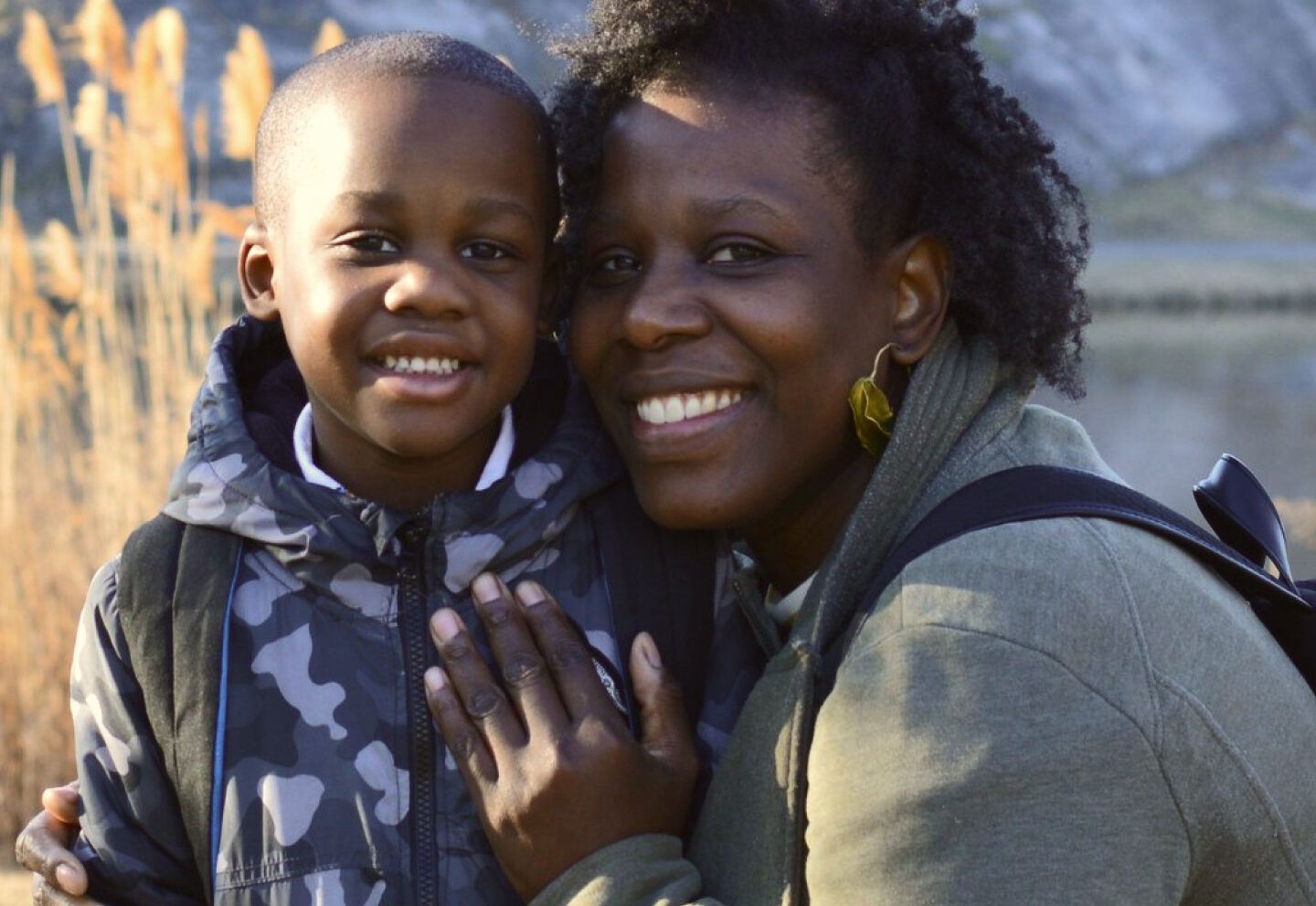 A Black mother and son are outside in the sunshine. The mother crouches to hug her son, their faces touching as they look at the camera, smiling.